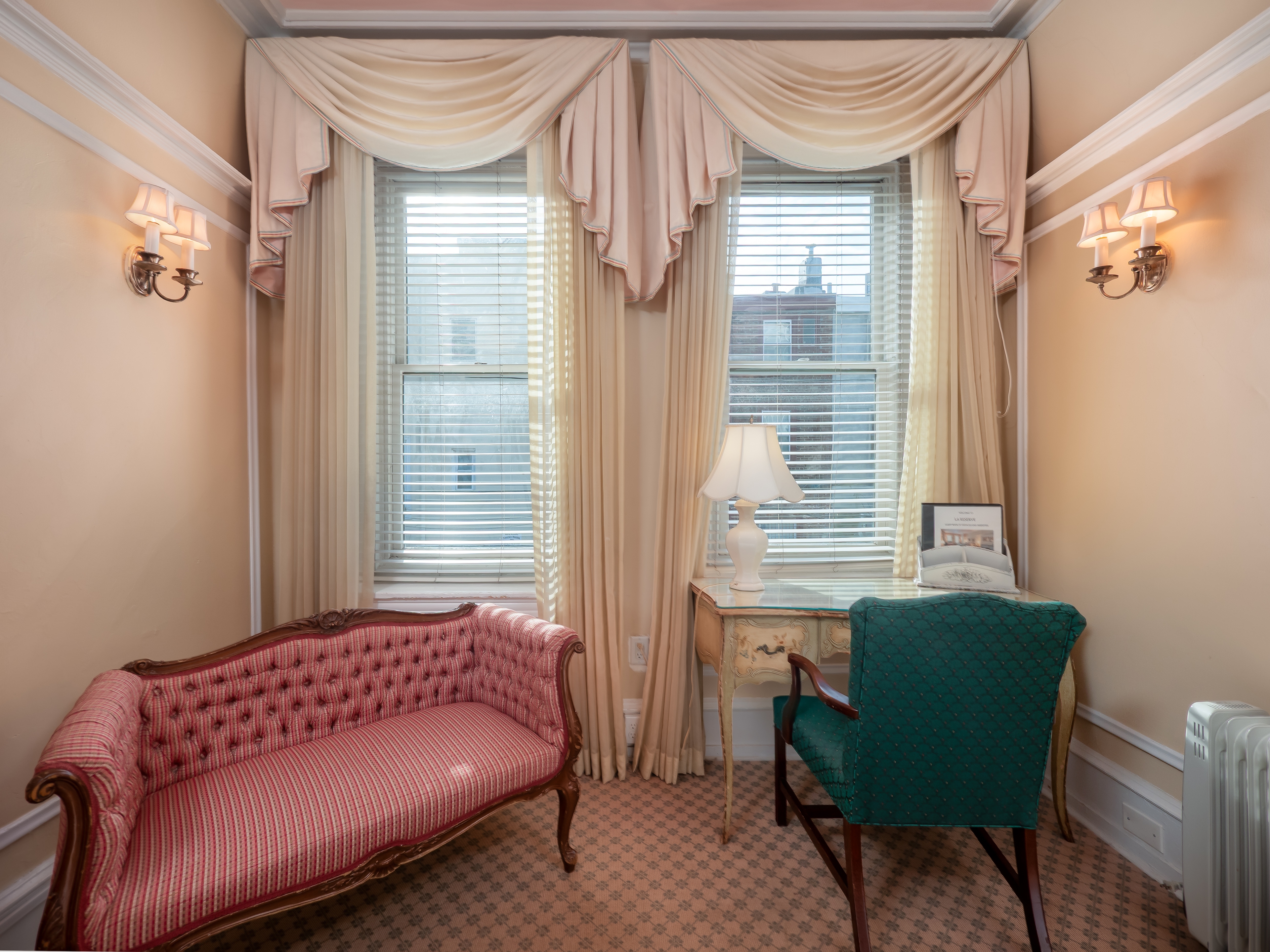 A vintage couch facing away from two windows with cushioned chair tucked in a desk facing one of the windows.