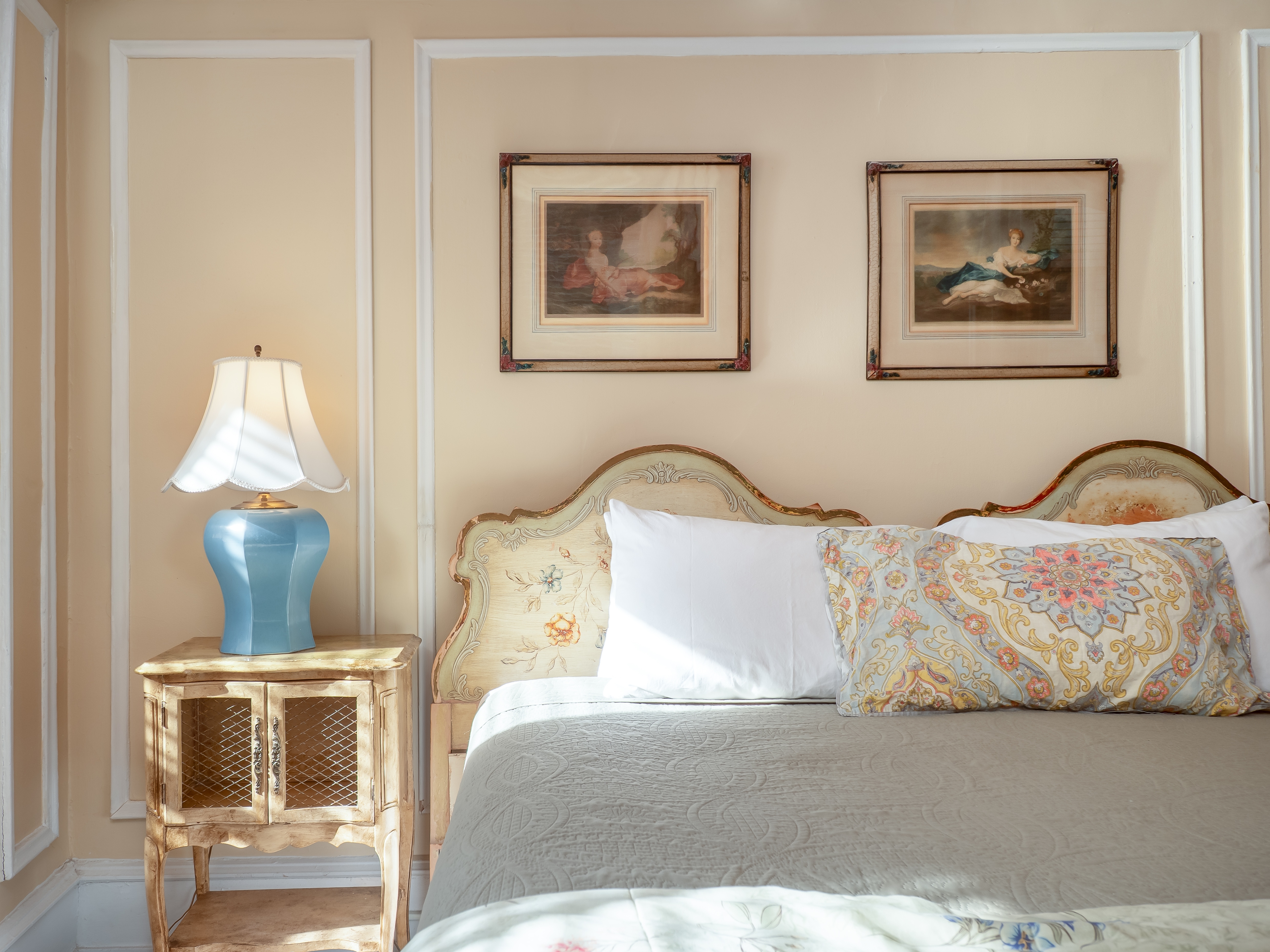 Close up of a queen bed, bedside table with an antique lamp and antique wall art.