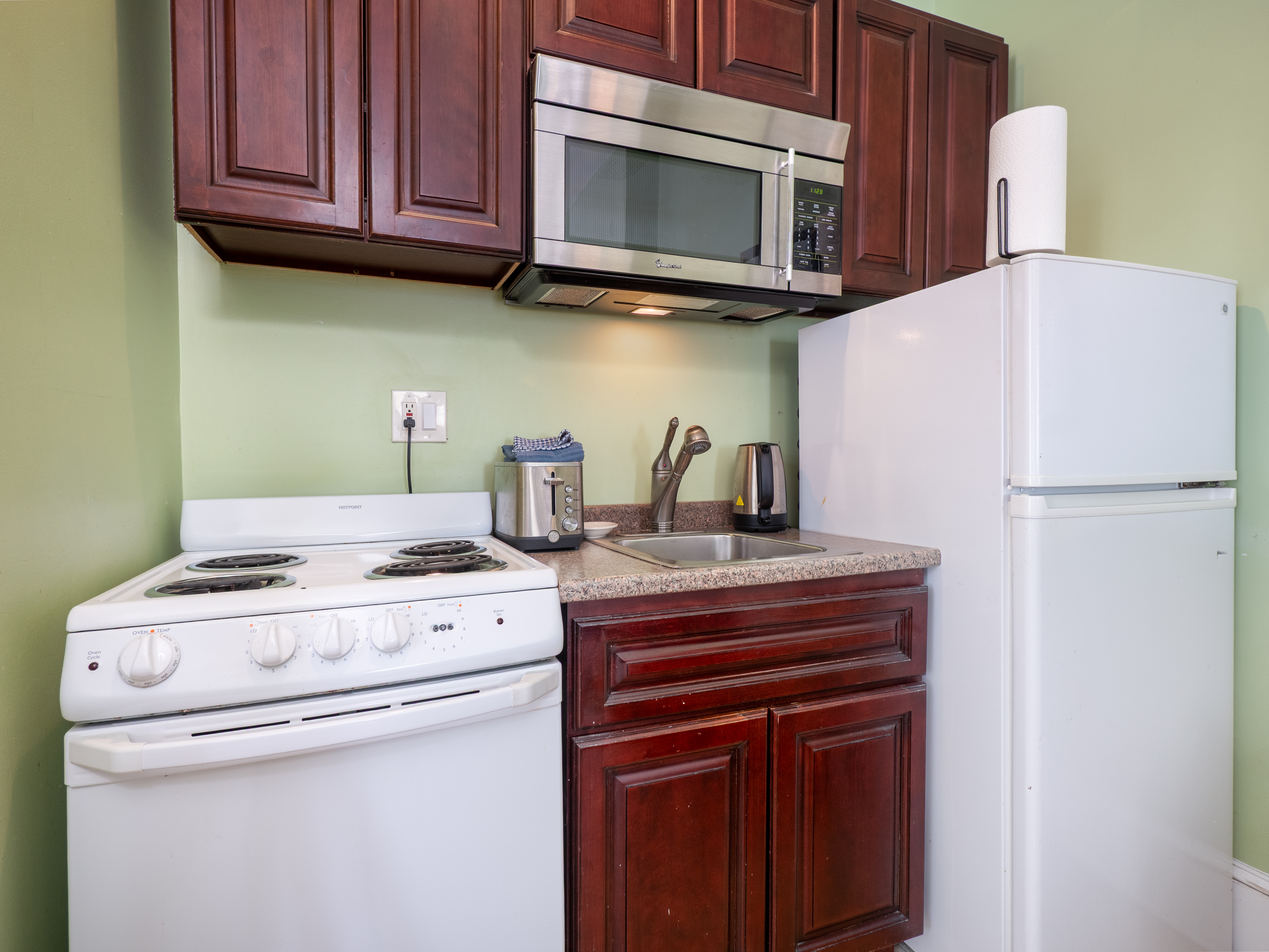 Closeup of the kitchenette that has a stove, sink, fridge, microwave and cabinets.