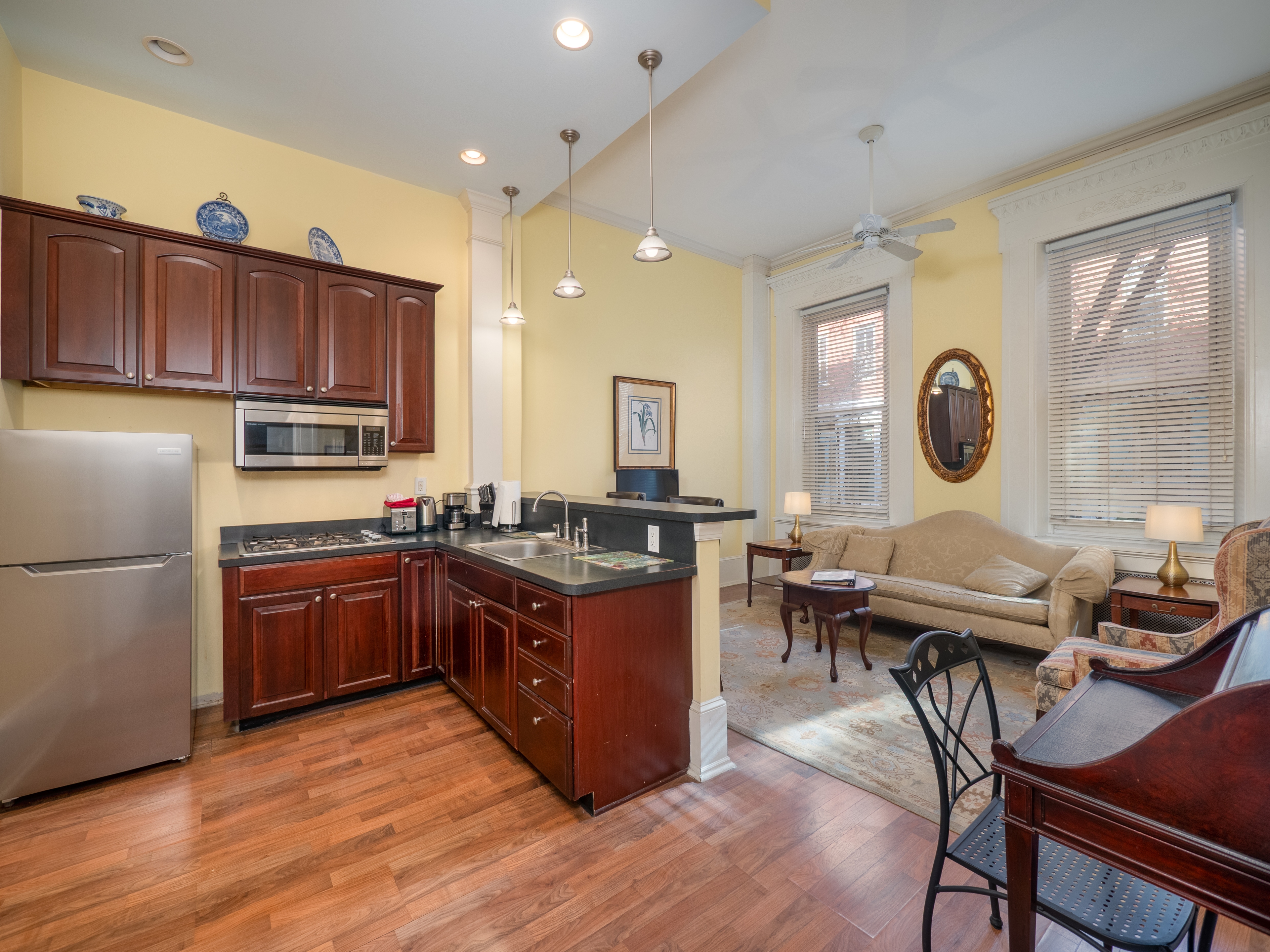 Full kitchen with sink, stove, fridge, microwave and cabinets. A sitting area with sofa, chairs and windows is behind.
