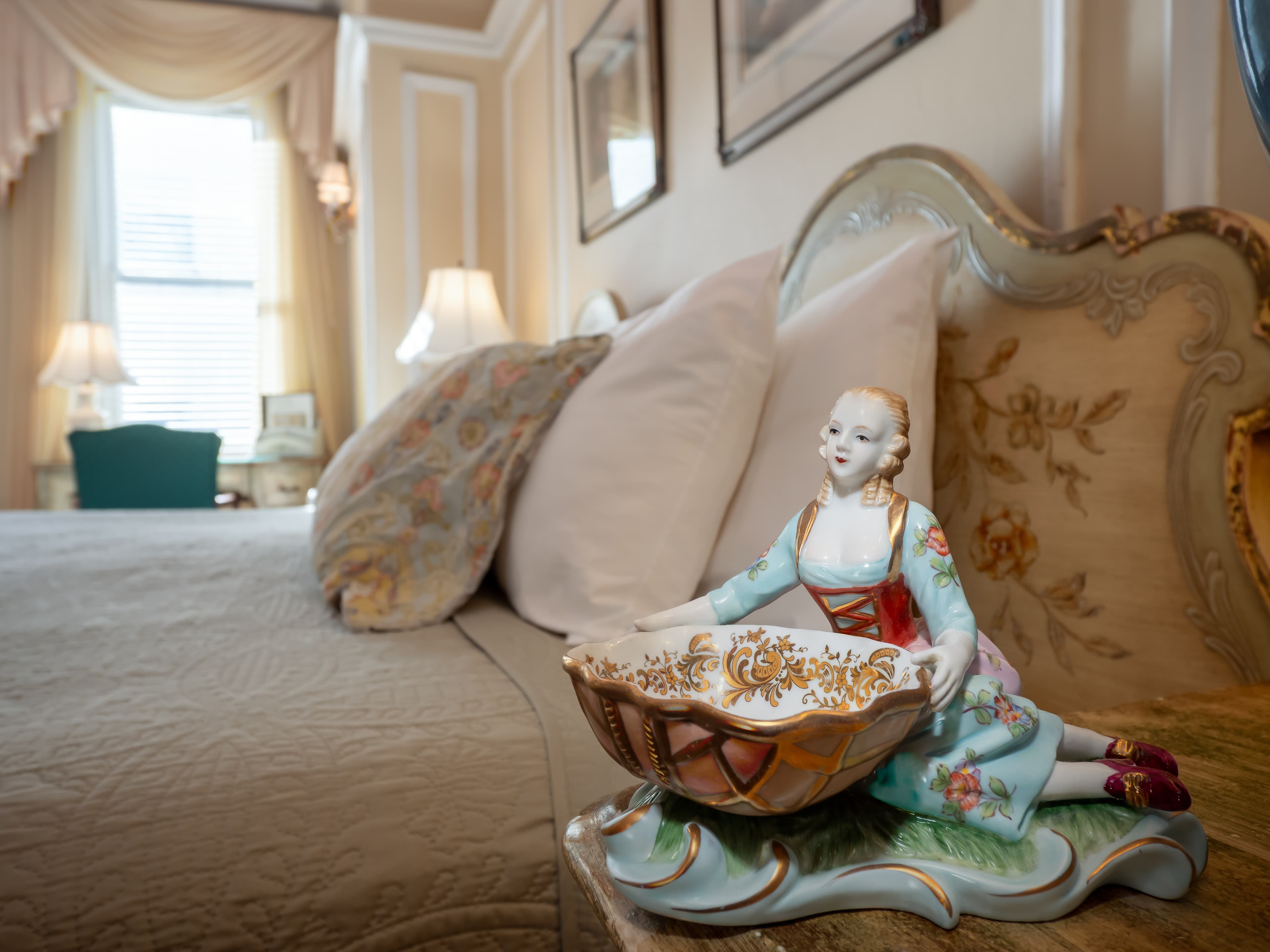 Close up of antique statuette with a queen sized bed and a window providing natural light in the background.