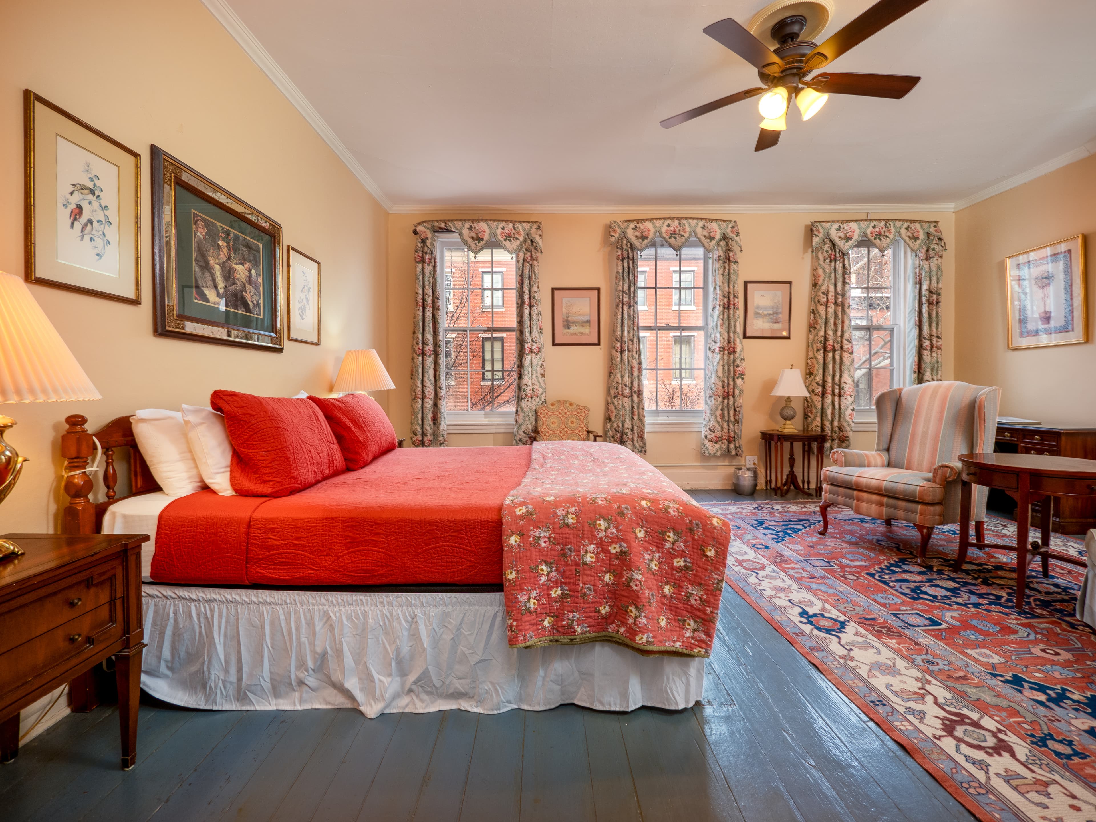 A cheery room with a queen bed, three windows, desk, and ceiling fan.