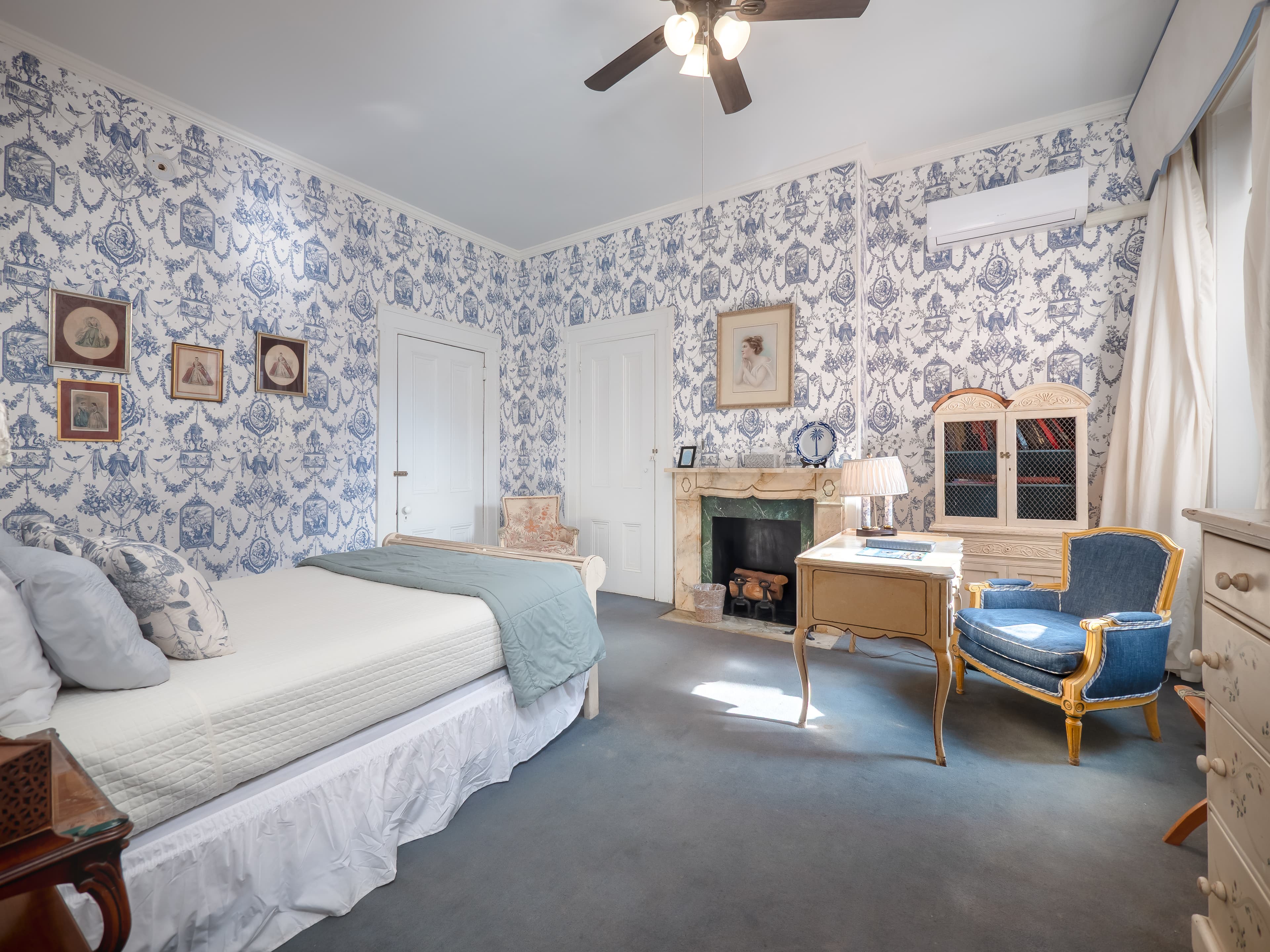 Elegant bedroom with a queen bed, decorative fireplace, ceiling fan, two cushioned chairs and windows.