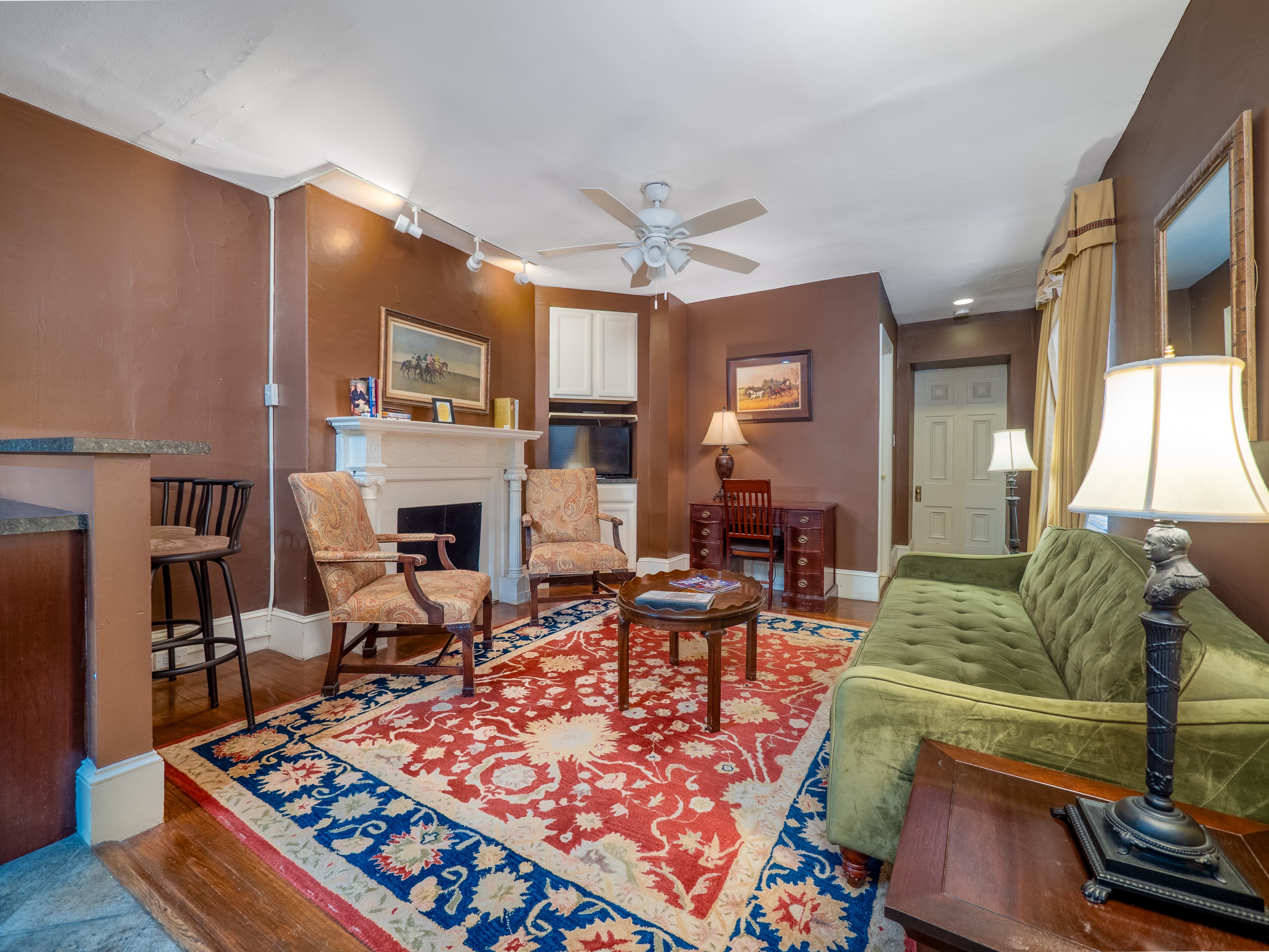 A homey living room with a couch, a mirror above the couch, two armchairs, a decorative fireplace, desk and TV.