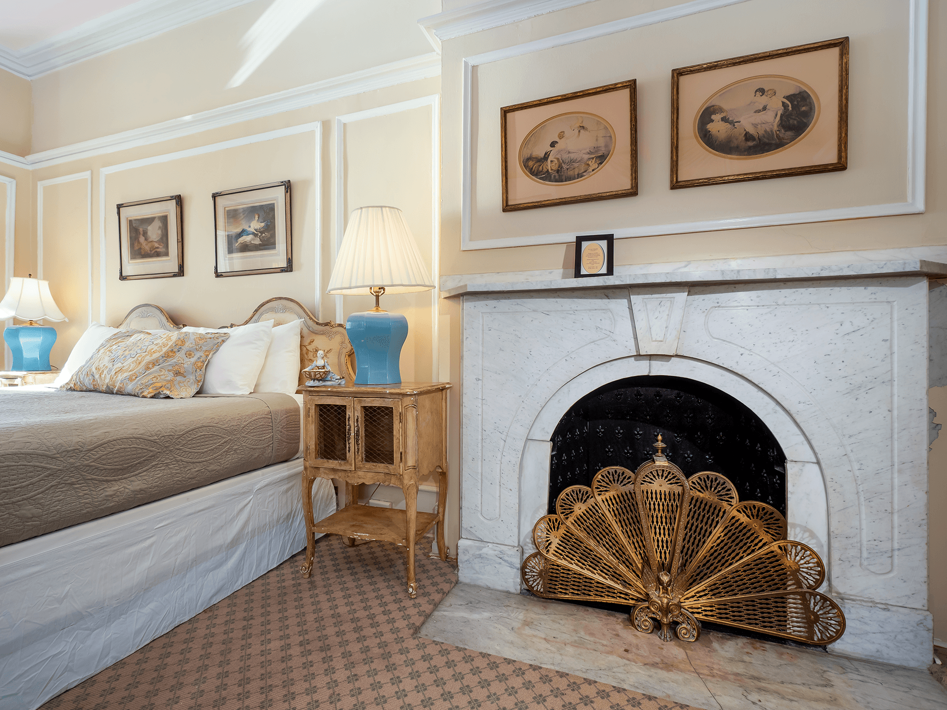 A cozy bedroom featuring a bed with decorative pillows, a lamp, a side table, and a marble fireplace with an ornate screen.