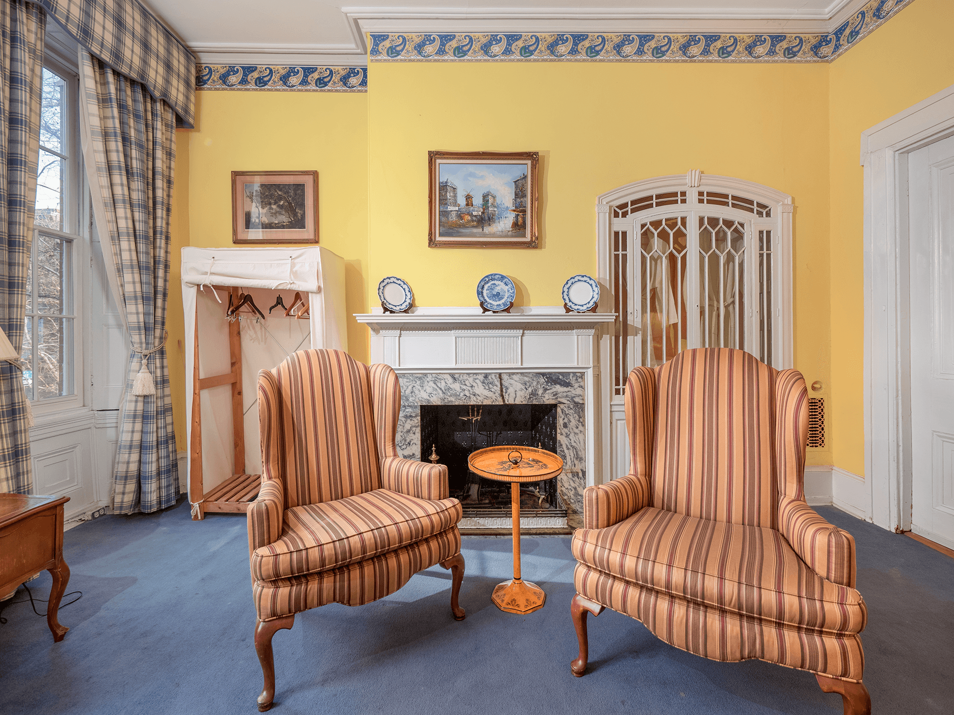 A cozy room featuring two striped armchairs, a small round table, a fireplace, and yellow walls adorned with artwork.