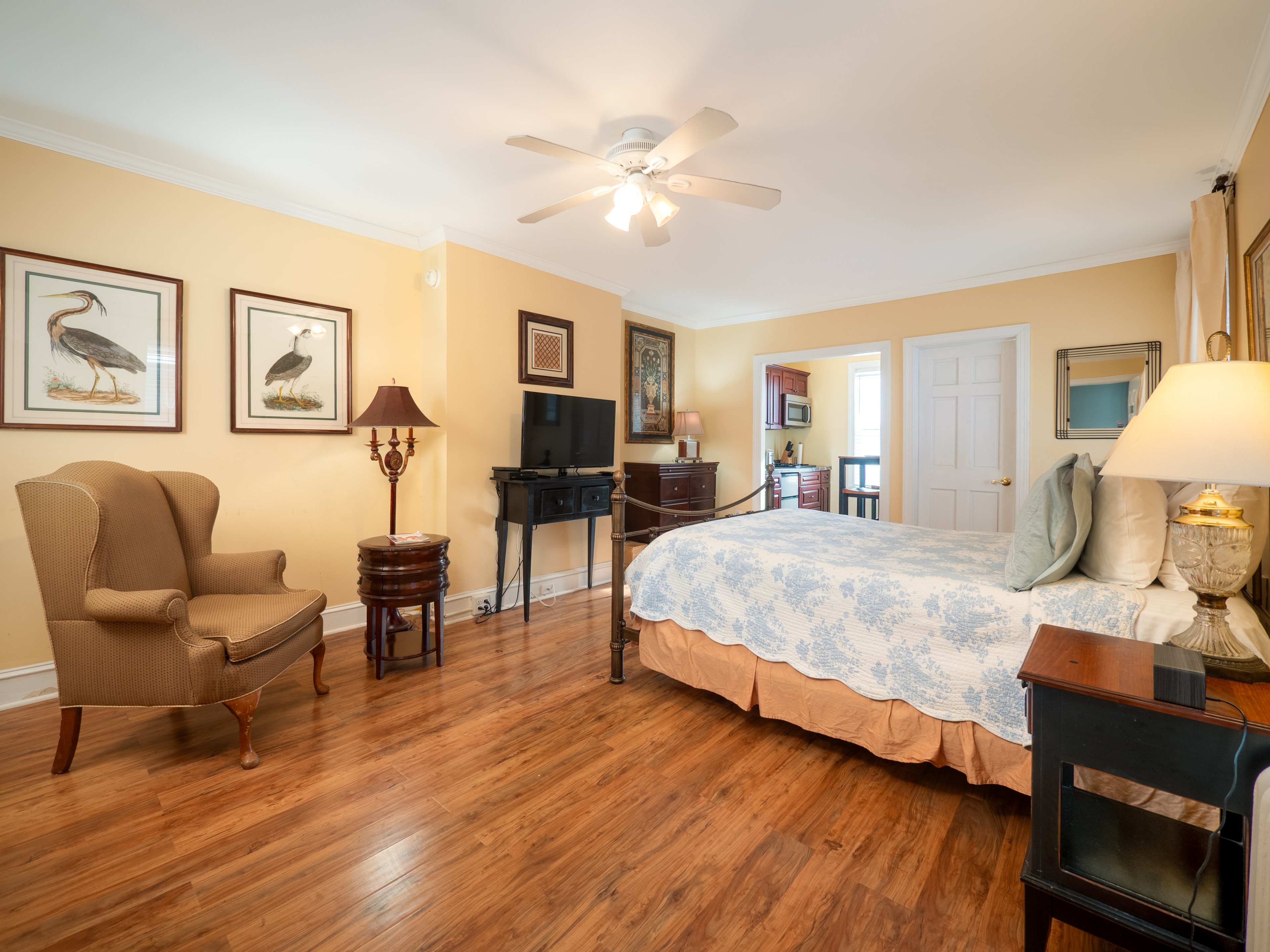 Sunny bedroom with a queen bed, TV across the bed, kitchenette, armchair and side table.