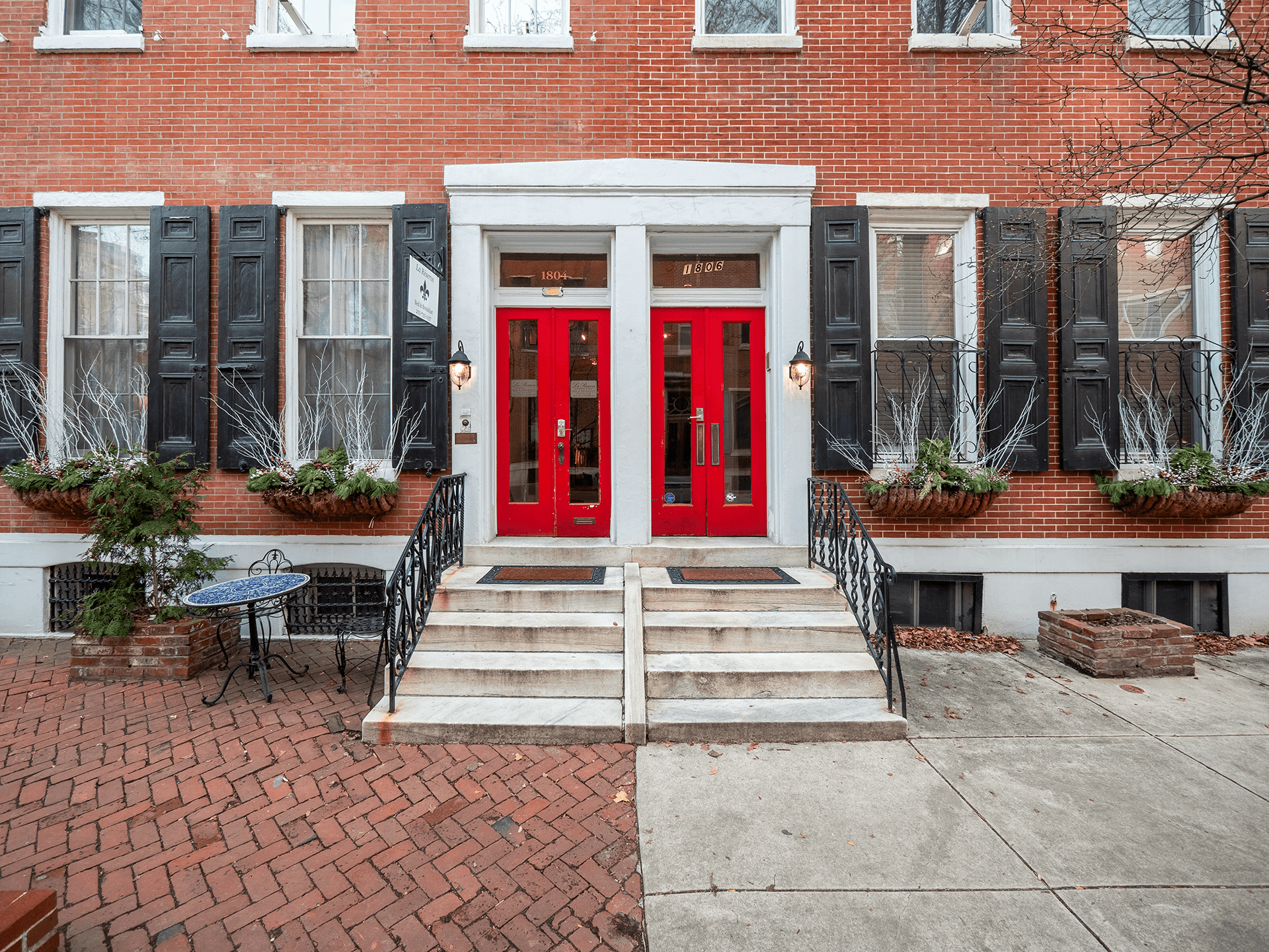 Entrance of a brick building featuring red doors, black shutters, and potted plants.