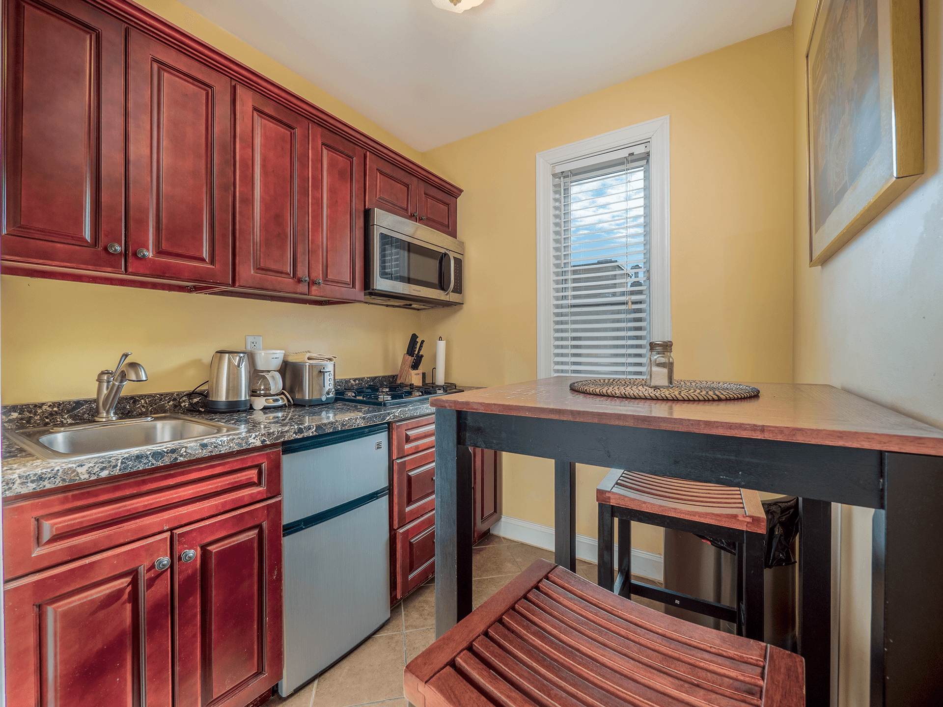 A small kitchen features red cabinets, stainless steel appliances, and a table with two stools under a bright window.